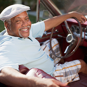 A cheerful senior man wearing a flat cap relaxes in the driver’s seat of a vintage convertible with a red leather interior.