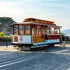 A classic wooden San Francisco cable car sits on tracks at a turnaround point under a clear sky, with trees and distant hills in the background.