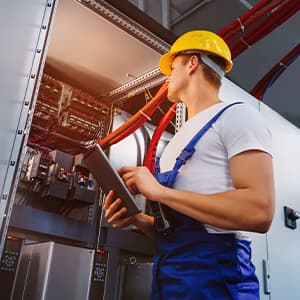 An electrician in a hard hat and protective workwear examines an open industrial electrical control cabinet while holding a tablet.