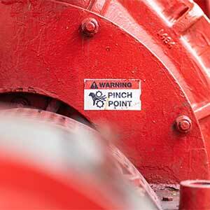 A close-up view of red industrial equipment shows a warning label that reads 'Pinch Point' with a hazard icon, indicating an area where moving machine parts could cause injury.