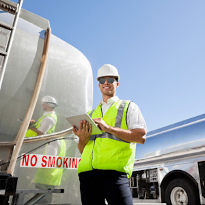 A smiling man wearing a hard hat and high visibility vest uses a tablet while inspecting gasoline tanker trucks.