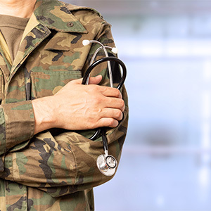 A person dressed in a camouflage military uniform crosses their arms while holding a stethoscope.