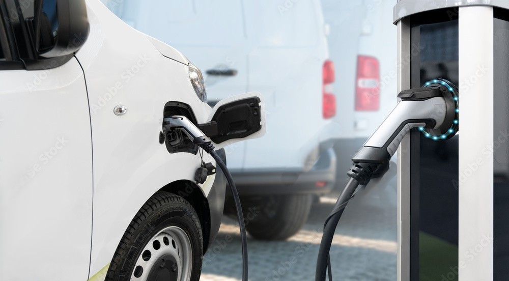 A white electric van plugged into a public EV charging station using a charging cable.