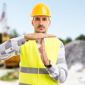 A male construction worker wearing a yellow hard hat and high-visibility safety vest holds his hands in a time-out gesture, signaling a stop or pause in work activity at an outdoor construction site.