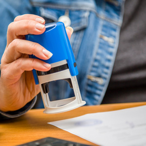A close-up of a person wearing a denim jacket pressing a blue self-inking notary stamp onto a paper document on a wooden desk.