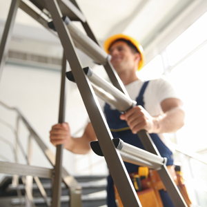 A worker in a yellow hard hat and tool belt climbs a ladder inside a building, with a stair railing visible in the background.