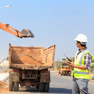 A road construction worker observes an excavator loading dirt into the bed of a dump truck.