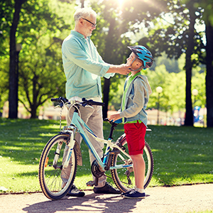 An adult adjusts a child’s bicycle helmet beside a bike before riding in a sunny park.