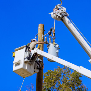 Utility worker repairing electrical equipment on power lines from bucket truck under a clear blue sky. 