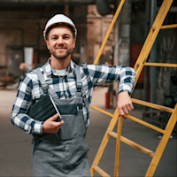 A smiling industrial worker wearing a white hard hat and protective overalls stands inside a manufacturing facility, leaning on a yellow ladder while holding a tablet.