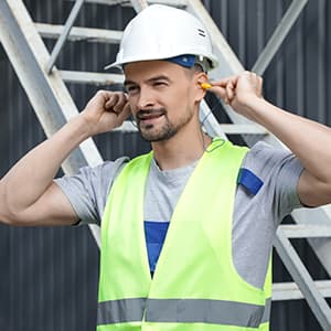A construction worker wearing a hard hat and high-visibility safety vest inserts earplugs to protect his hearing at an industrial worksite.