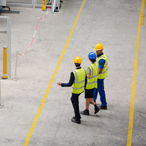 Three supervisors in high-visibility vests and hard hats walk between yellow aisle lines in a factory or warehouse.