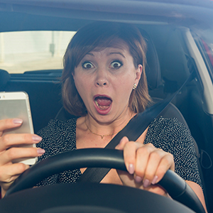 A woman behind the wheel of a car looks shocked and holds a smartphone in one hand.