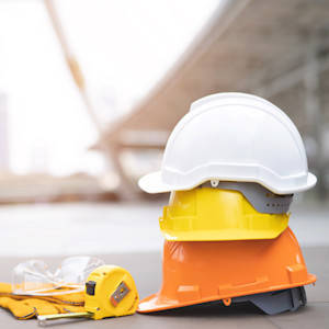 Three stacked safety helmets in white, yellow and orange sit on a flat surface beside yellow work gloves, clear safety glasses and a yellow tape measure, with a construction site blurred in the background.