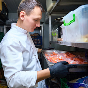A caucasian male kitchen employee places a tray of raw salmon on a shelf in a walk-in fridge.
