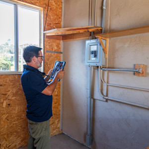 Middle-aged white male electrical inspector looks at a tablet while inspecting an electrical panel in a home under construction.