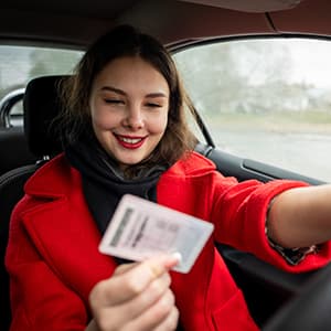 A woman wearing a red coat sits in the driver’s seat and holds up a driver’s license while keeping her other hand on the steering wheel.