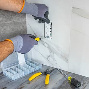 A gloved electrician uses a screwdriver to install an electrical outlet in a tiled wall, with tools and hardware organized on the countertop below.