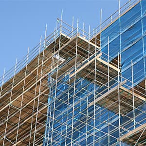 Metal scaffolding with protective netting is erected around a multi-story building under construction, providing elevated platforms and fall protection for workers during exterior work.