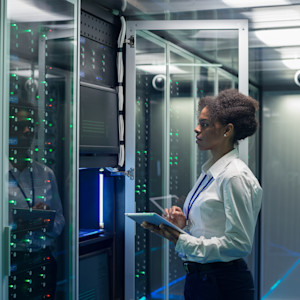A black female technician holding a tablet examines a server in a data center.