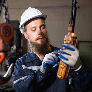 A bearded industrial worker wearing a white hard hat and blue protective gloves carefully examines a crane hoist inside a manufacturing facility.