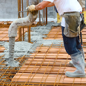 A construction worker wearing rubber boots and gloves pours wet concrete from a hose onto a reinforced steel rebar grid.