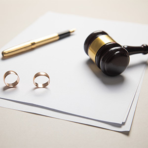 Two gold wedding rings lie on a stack of official notary documents, accompanied by a judge’s gavel and a black-and-gold pen. 