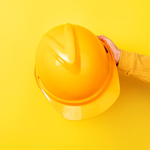 A close-up studio shot shows a person’s hand in a yellow sleeve holding a glossy yellow safety hard hat against a solid yellow background.