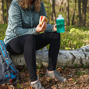 A woman rests on a fallen log in a wooded area while holding a sandwich and a reusable water bottle beside her backpack.
