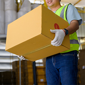 A warehouse worker wearing a reflective safety vest and gloves carefully lifts a large cardboard box. The background shows industrial shelving and stacked boxes.