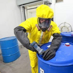 A worker wearing a yellow hazmat suit, full-face respirator and heavy protective gloves seals the lid of a blue chemical drum inside a controlled storage area. 