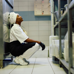 An exhausted black female professional cook crouches next to shelves in a commercial kitchen.