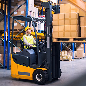 A warehouse employee wearing a yellow safety vest and hard hat operates a powered industrial truck, carefully lifting a pallet of stacked cardboard boxes in an organized storage facility.