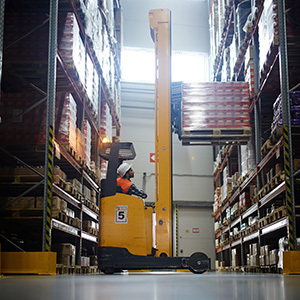 A warehouse worker operates a reach truck between tall storage racks while lifting a pallet, dmeonstrating safe powered industrial truck use in a warehouse setting.