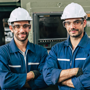 Two male factory workers wearing white hard hats, clear safety goggles and blue coveralls stand confidently with arms crossed in front of industrial machinery in a manufacturing facility.