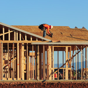 A construction worker wearing an orange safety shirt and helmet is positioned on the roof, securing plywood sheathing onto a wooden house frame. 