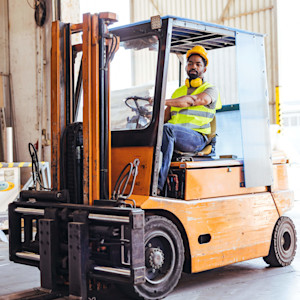 A warehouse worker wearing a hard hat, safety vest and hearing protection sits on an orange forklift near stacked materials in an industrial storage area.