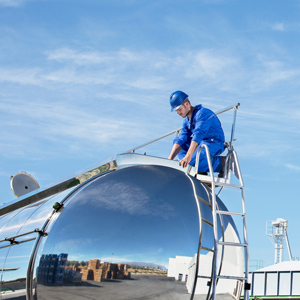 A worker wearing blue coveralls and a hard hat climbs a ladder attached to a large storage tank at an industrial site, with a clear blue sky in the background.