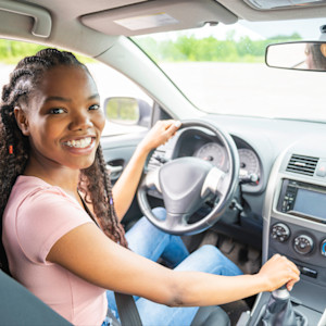 A smiling black female teenage driver sitting in the driver's seat of her car.