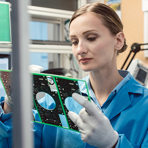 A technician wearing protective gloves and a blue lab coat carefully examines electronic circuit boards in a laboratory.