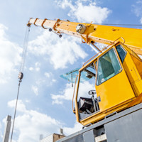 A large yellow mobile crane extends its boom high into a bright blue sky as it hoists a load on steel cables, with the operator cab visible and construction structures in the background.