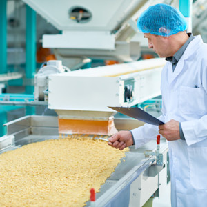 A food processing facility worker inspects a conveyor belt carrying dried pasta noodles in a food production plant.
