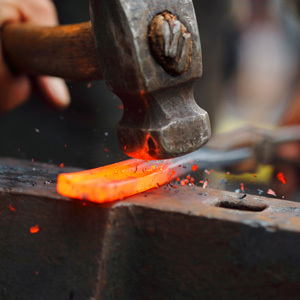 A blacksmith strikes a piece of heated metal with a hammer on an anvil, causing bright orange sparks to scatter. 