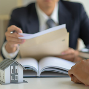 A real estate agent in a suit reviews paperwork with a client at a desk, with a small model house and an open book in the foreground.