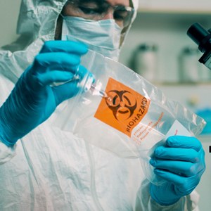 A lab worker wearing gloves, a mask and protective clothing examines a clear biohazard bag in a laboratory setting.