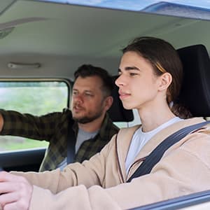 A driving instructor sits beside a young person in the driver’s seat, pointing ahead as he provides driving guidance inside a car.