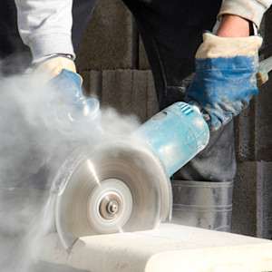 A worker wearing protective gloves operates a handheld power saw to cut through a concrete block, with fine dust billowing into the air.