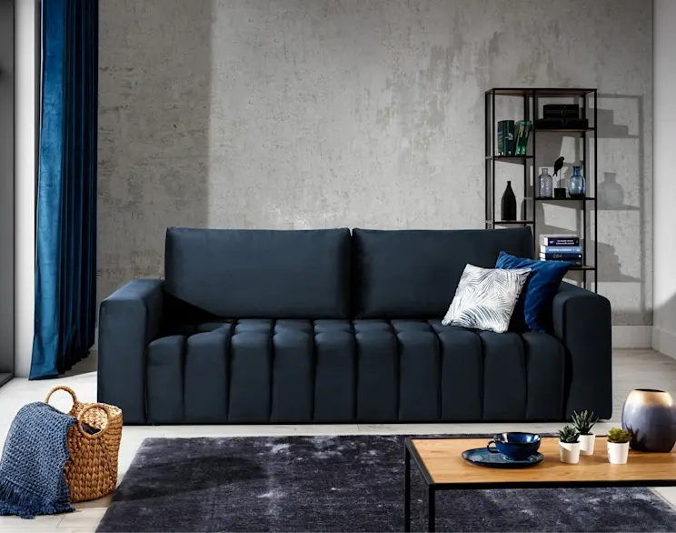 Granatowa kanapa Lazaro Monolith 79 z pionowym pikowaniem, w nowoczesnym salonie na tle betonowej ściany, z stolikiem i dodatkami.