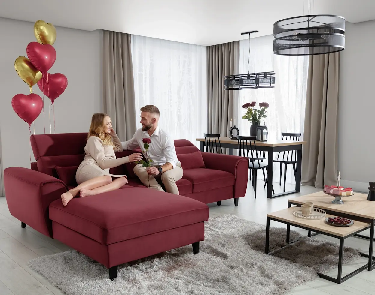 Walentynkowa aranżacja salonu z czerwonym narożnikiem Noble i dekoracjami w kształcie serc. Para odpoczywa na sofie, a jasne wnętrze z dużymi oknami i stolikiem kawowym tworzy romantyczny klimat.