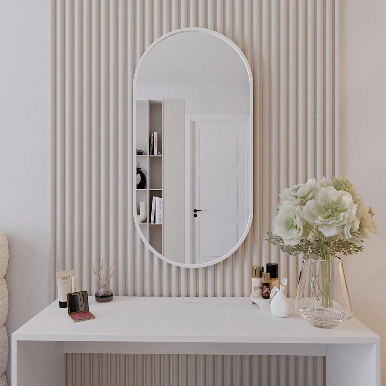 Białe, owalne lustro Arducati zawieszone na ścianie z pionowymi lamelami w jasnym kolorze. Pod spodem stoi białe biurko Nevy z dekoracjami: kosmetykami, dyfuzorem zapachowym i wazonem z jasnymi kwiatami.